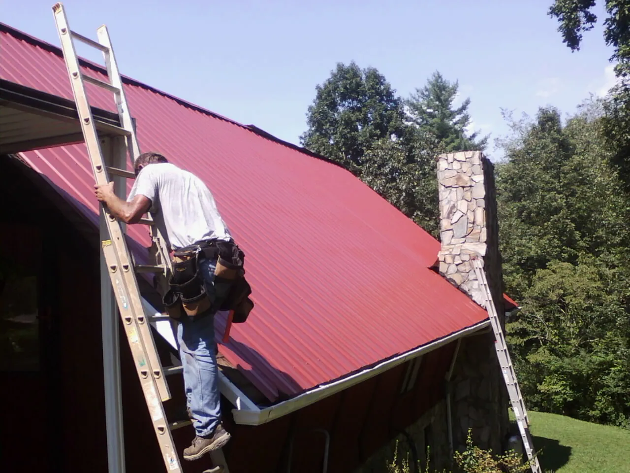 Roofer on a ladder beside a red metal roof and stone chimney — Soffit & Fascia Repair by Roofing Brothers in Eldridge